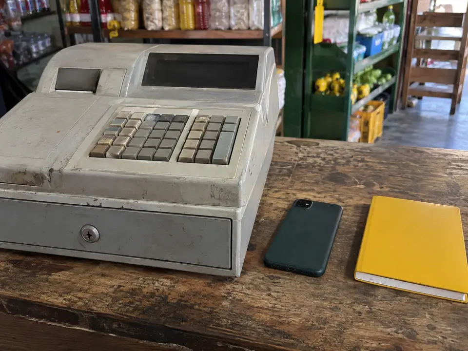 Caja registradora gris y gastada junto a un celular boca abajo y una libreta amarilla