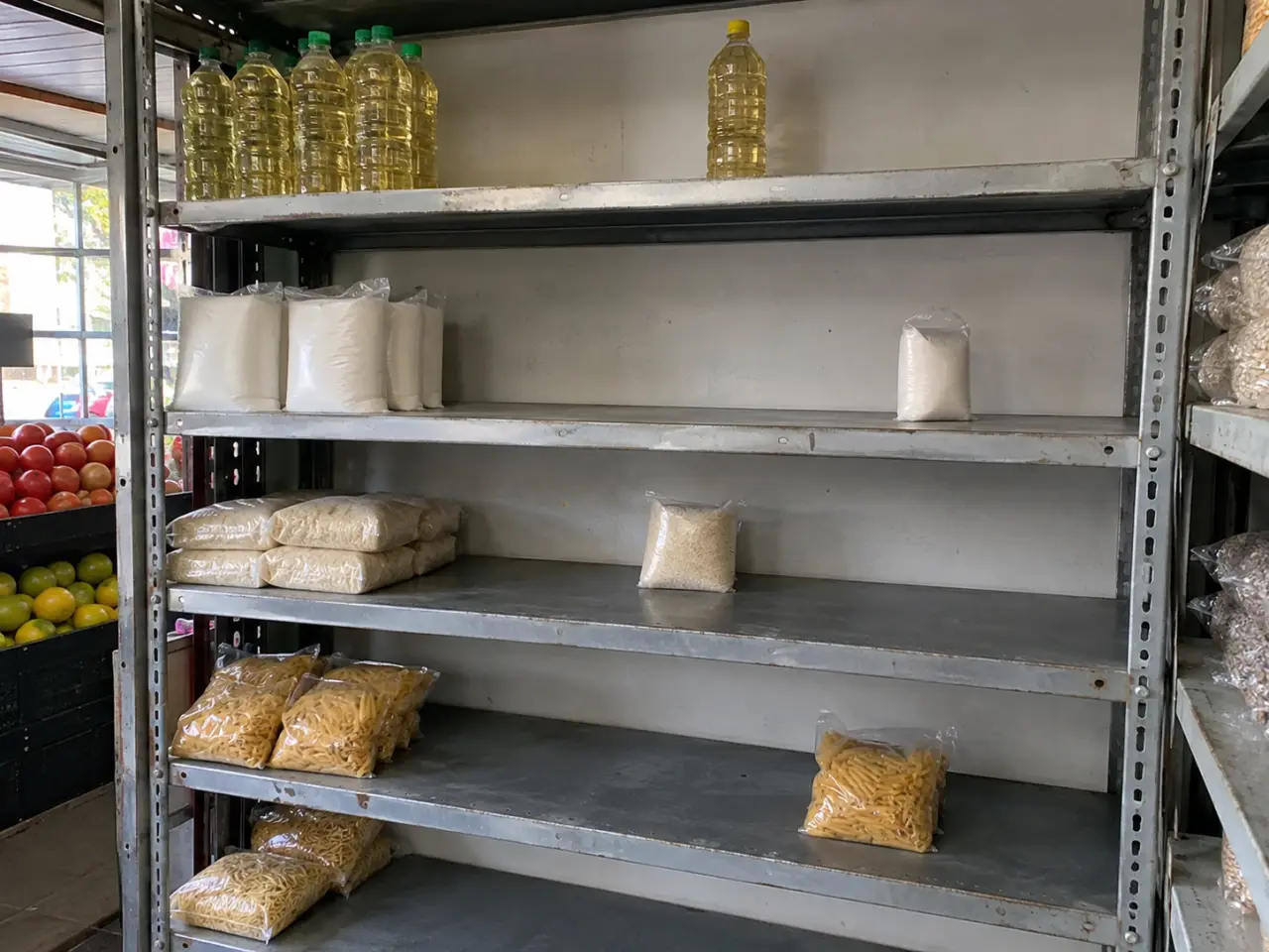 Estante metálico con aceite, azúcar, arroz y fideos, varias filas casi vacías