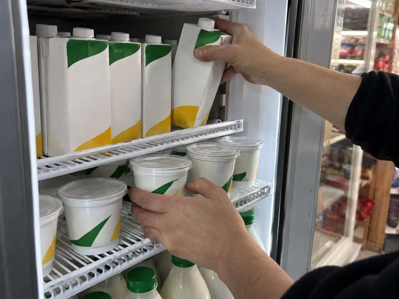 Manos reponiendo cajas de leche y potes de yogur en el refrigerador vertical de un almacén