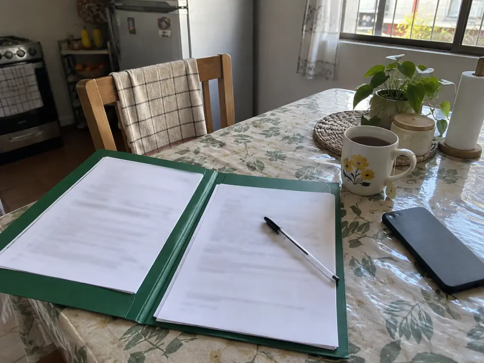 Carpeta verde con hojas, lápiz, taza de té y celular sobre una mesa de cocina con hule floreado